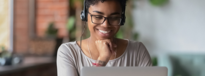 A black feminine person is using a laptop, she is wearing a headset and is resting her head on her hand whilst smiling. 