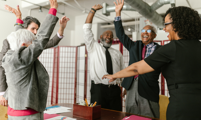 A group of older people in smart attire of different races and gender presentation throw their hands joyfully in the air while they're laughing.