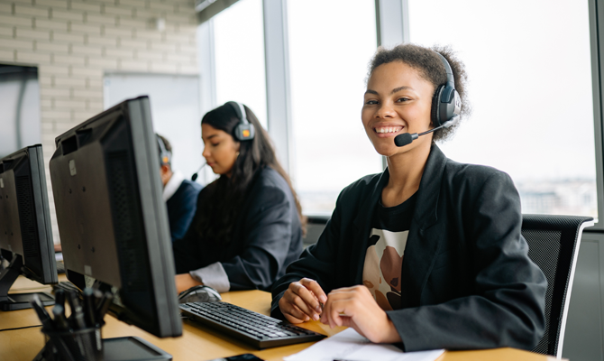 A young black feminine person sits at a call centre desk in a blazer with headphones on and smiles at the camera happily.