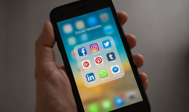 A white hand holds a phone showing social media icons in a folder call social networks. 