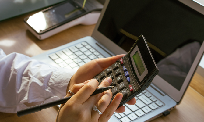 A wooden desk with a laptop on, someone with a calculator is calculating something.