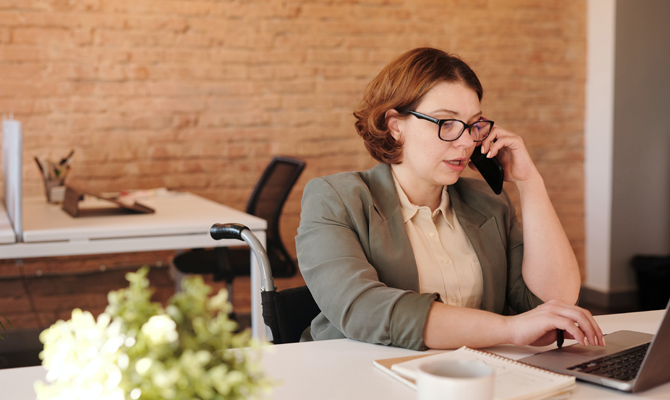 to represent how to reduce customer service costs a feminine white person sits at a laptop on the phone. They are a wheelchair user, wearing a blazer in a brick office.