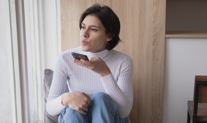 A light skinned person with short brown hair talks into a phone. They're wearing a ribbed white long sleeved top, blue jeans and sitting in a light room with wood and white walls.