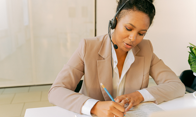 A black feminine person wears a beautiful camel colour blazer, a call centre headset and writes on a desk.